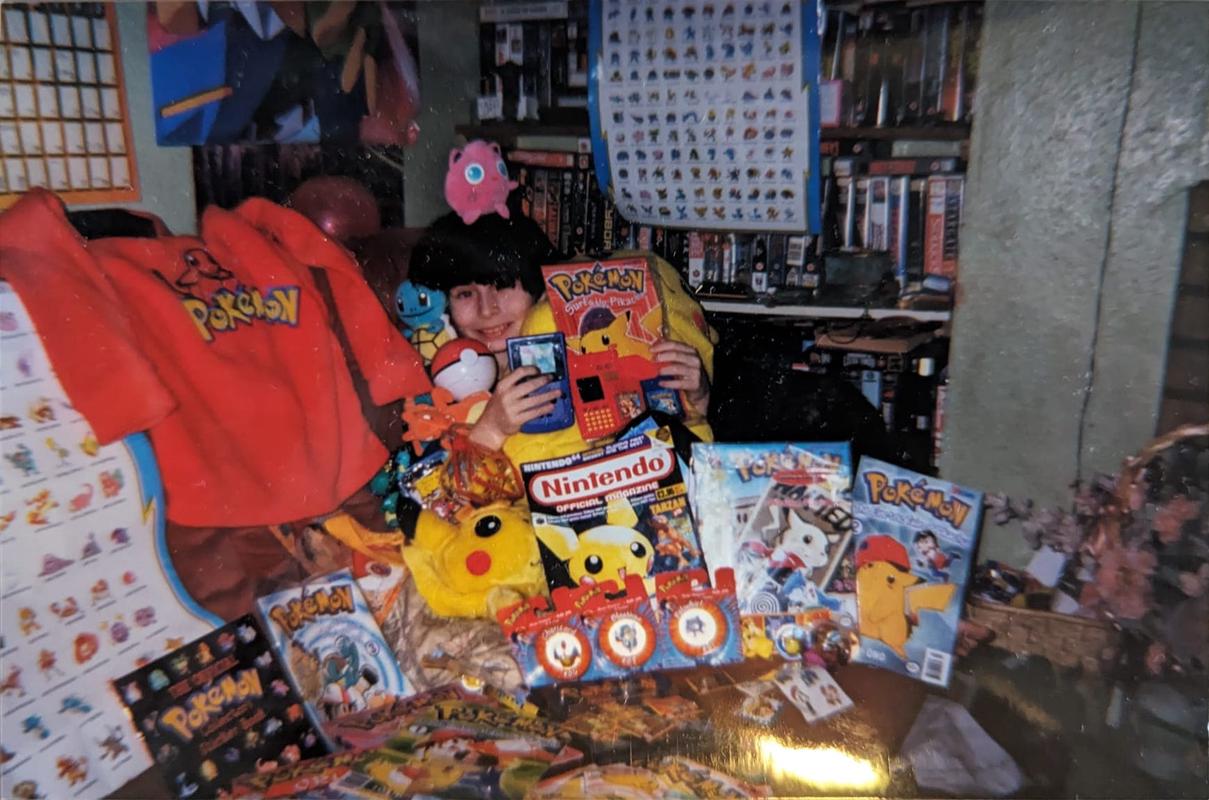 A photograph of Dazz, who appears to be drowning in Pokémon toys, magazines, games, and other Pokémon items. Any suggestion that he was a massive Pokémon fan as a kid, would be an understatement!